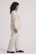 A model stands against a light gray studio background wearing a cream-colored hemp blazer with a yellow and brown eco-print, a mustard yellow collared shirt, cream wide-leg trousers, and black shoes.