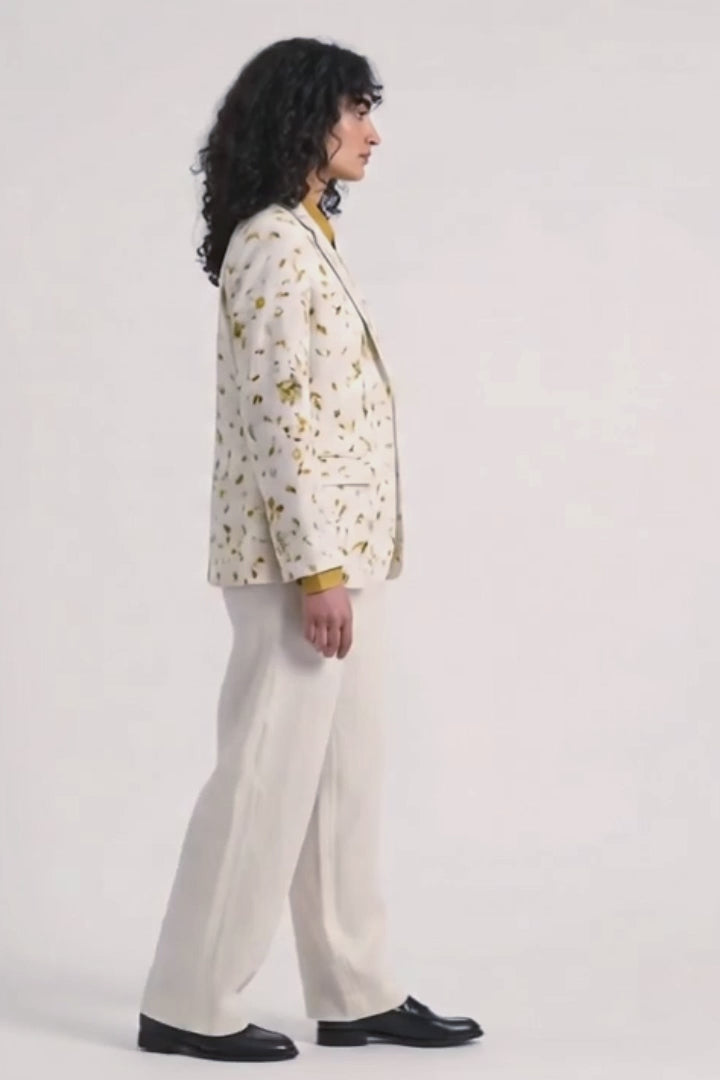 A model stands against a light gray studio background wearing a cream-colored hemp blazer with a yellow and brown eco-print, a mustard yellow collared shirt, cream wide-leg trousers, and black shoes.