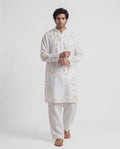A man modeling a white long-sleeve kurta with a subtle gold floral pattern, paired with white trousers and brown sandals, against a plain white studio background.