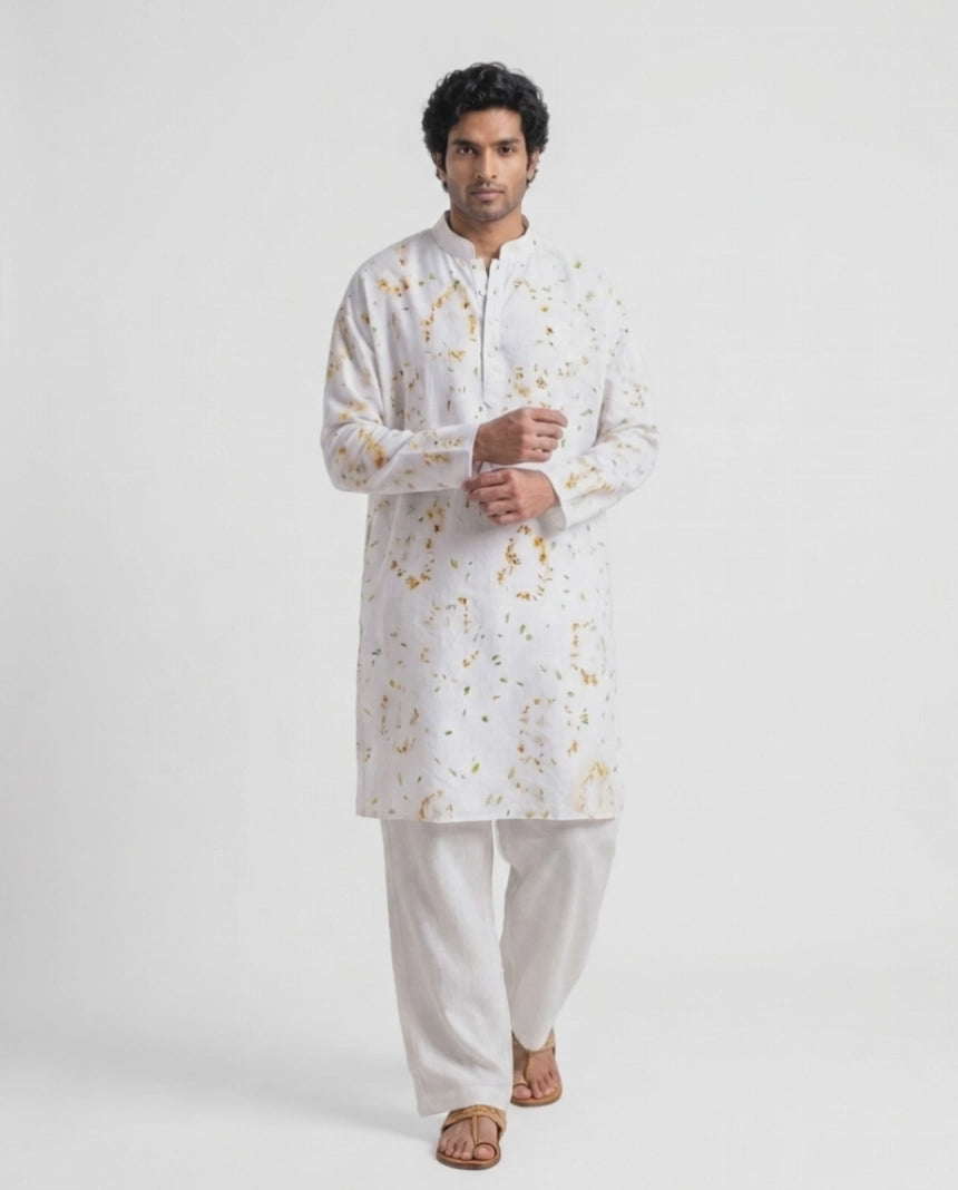A man modeling a white long-sleeve kurta with a subtle gold floral pattern, paired with white trousers and brown sandals, against a plain white studio background.