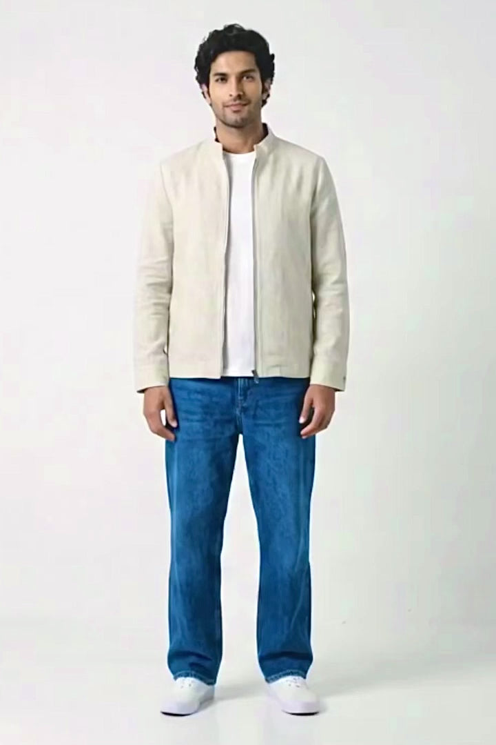A man wearing a light beige linen jacket with a stand collar and front zipper, paired with blue jeans and white sneakers, standing against a white studio background.